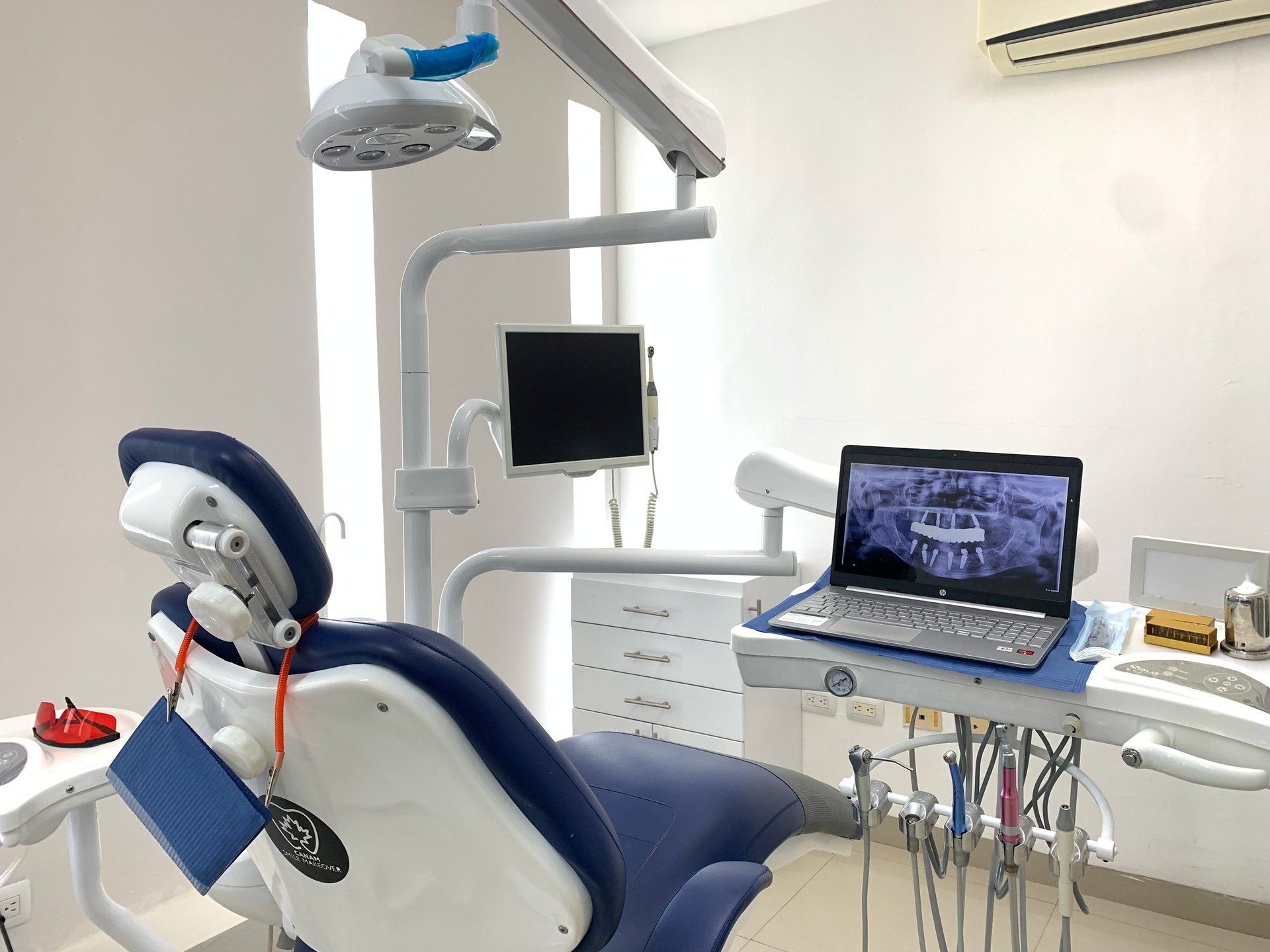 Modern dental treatment room with blue chair, overhead surgical light, monitor screens, and white cabinetry