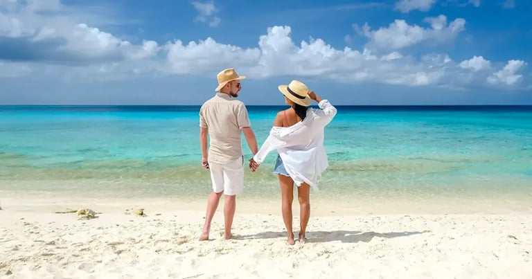 Couple in white clothing and hats standing on a sandy beach facing turquoise ocean water under blue sky