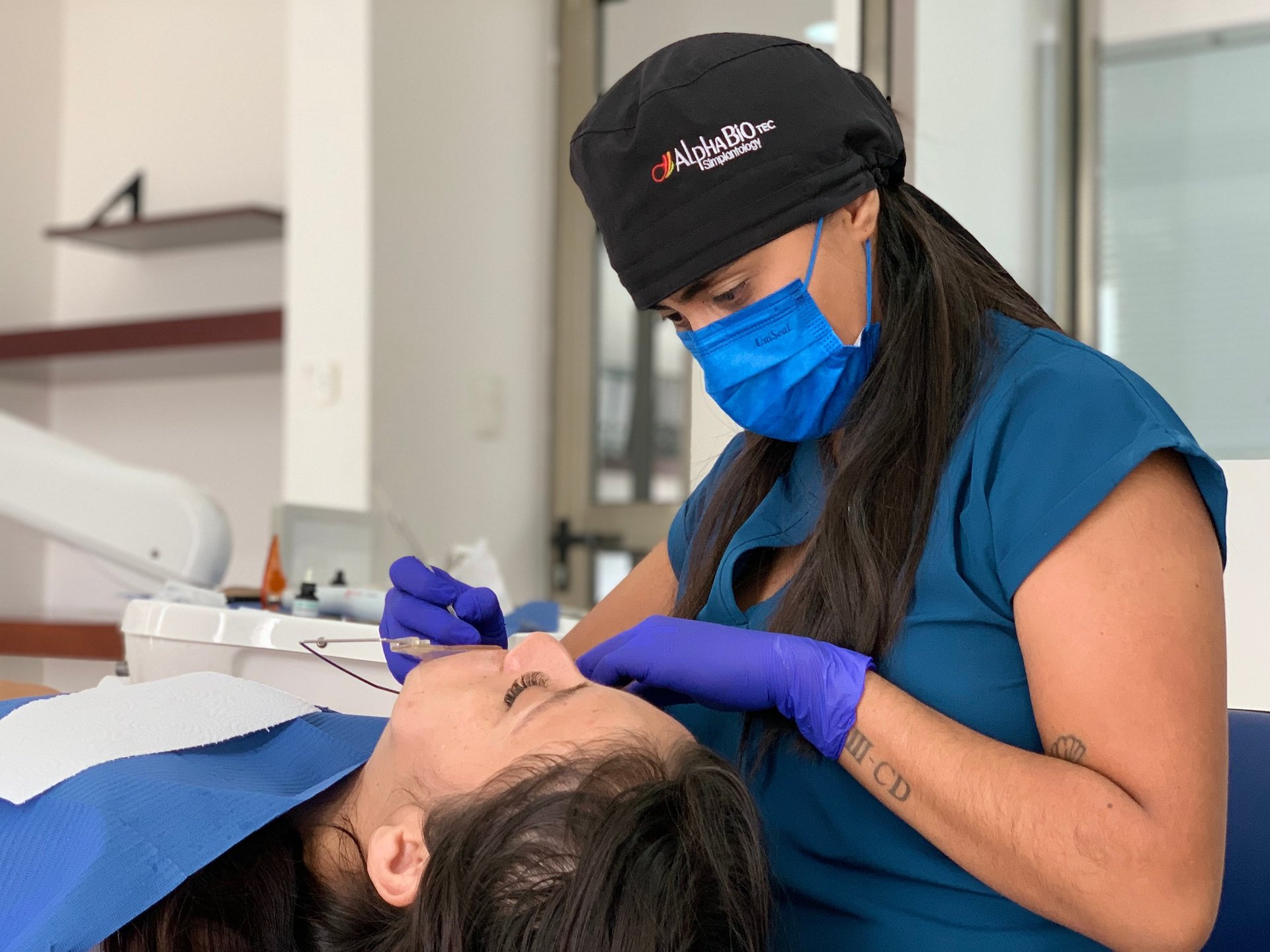 Female dental professional wearing blue scrubs, mask, and cap performing a dental procedure on a patient in a clinical setting