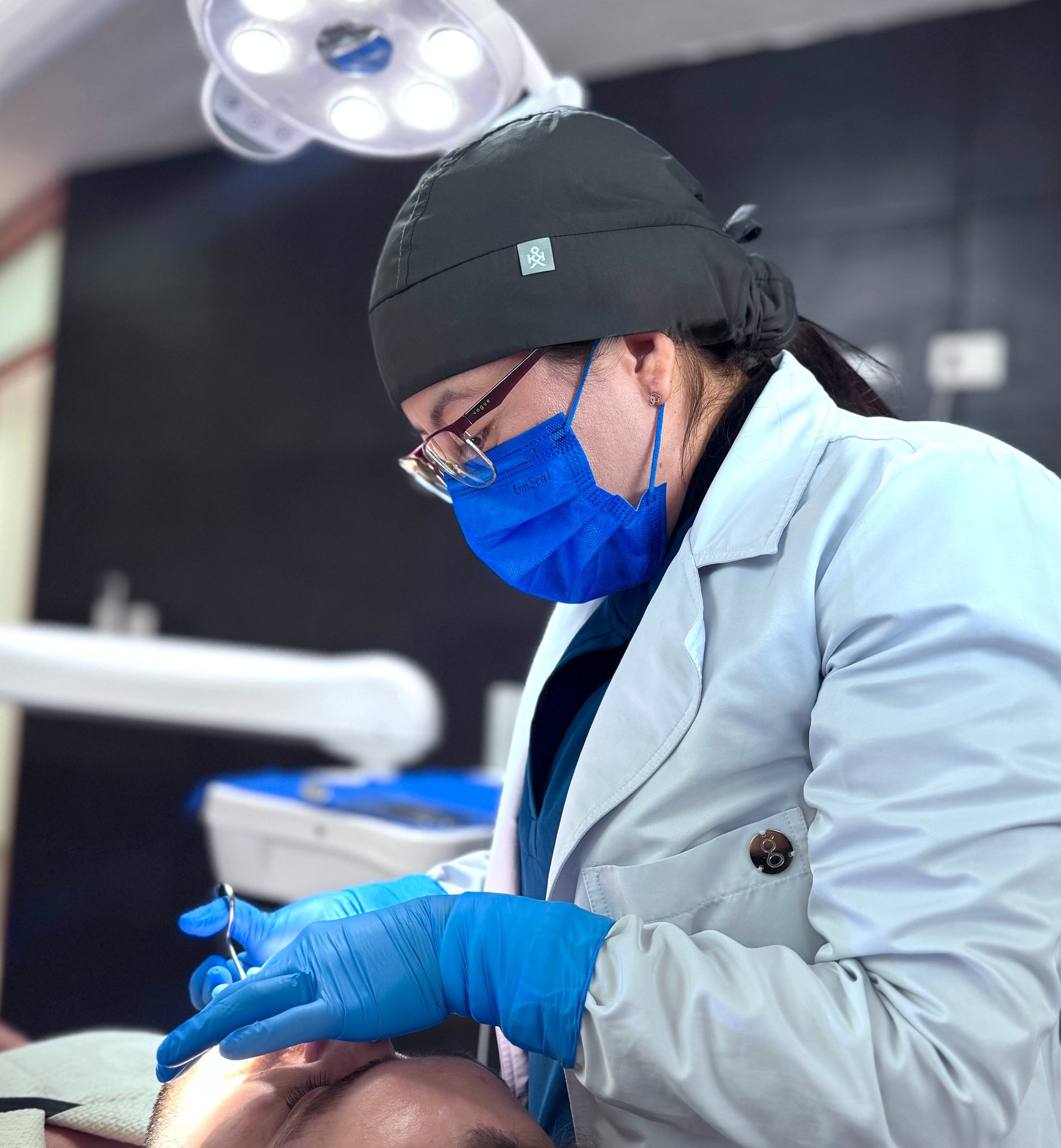 Dentist in surgical mask, headlamp, and blue gloves examining a patient in a dental office setting